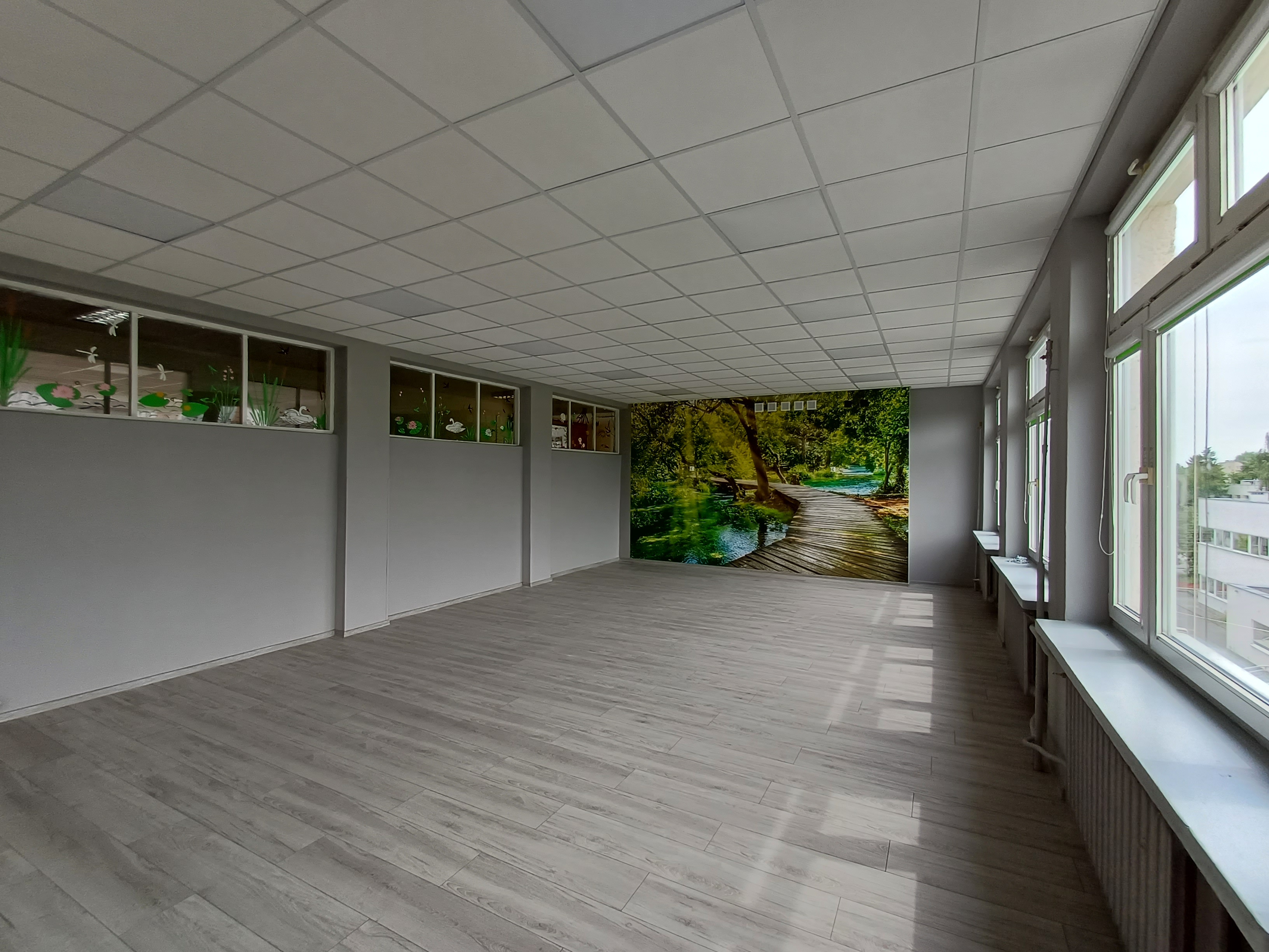 Odświeżona sala z nowym sufitem podwieszanym, panelami podłogowymi oraz fototapetą.